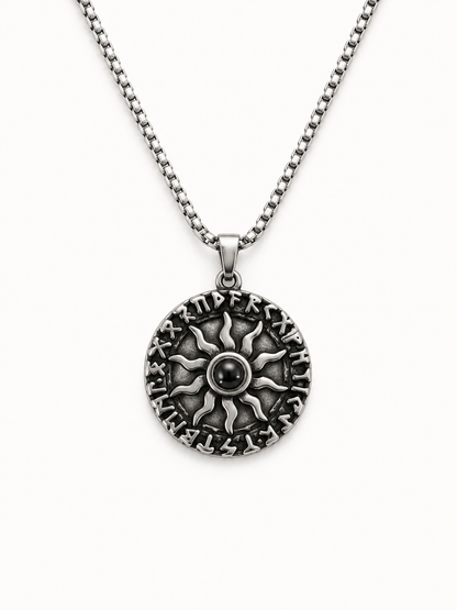 A silver-toned necklace featuring a circular pendant with an embossed sunburst design and raised lettering around the edge, suspended from a matching box chain against a plain white background.