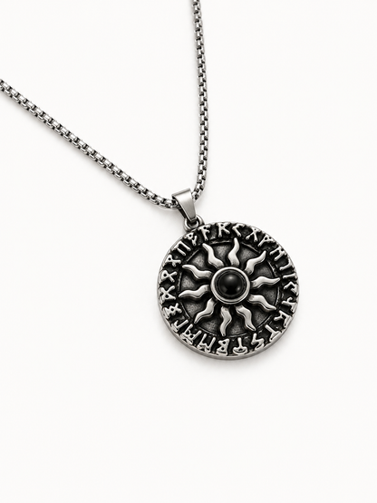 A silver-toned round pendant necklace featuring a sunburst design with engraved details and a central black bead, suspended from a matching chain on a light gray background.