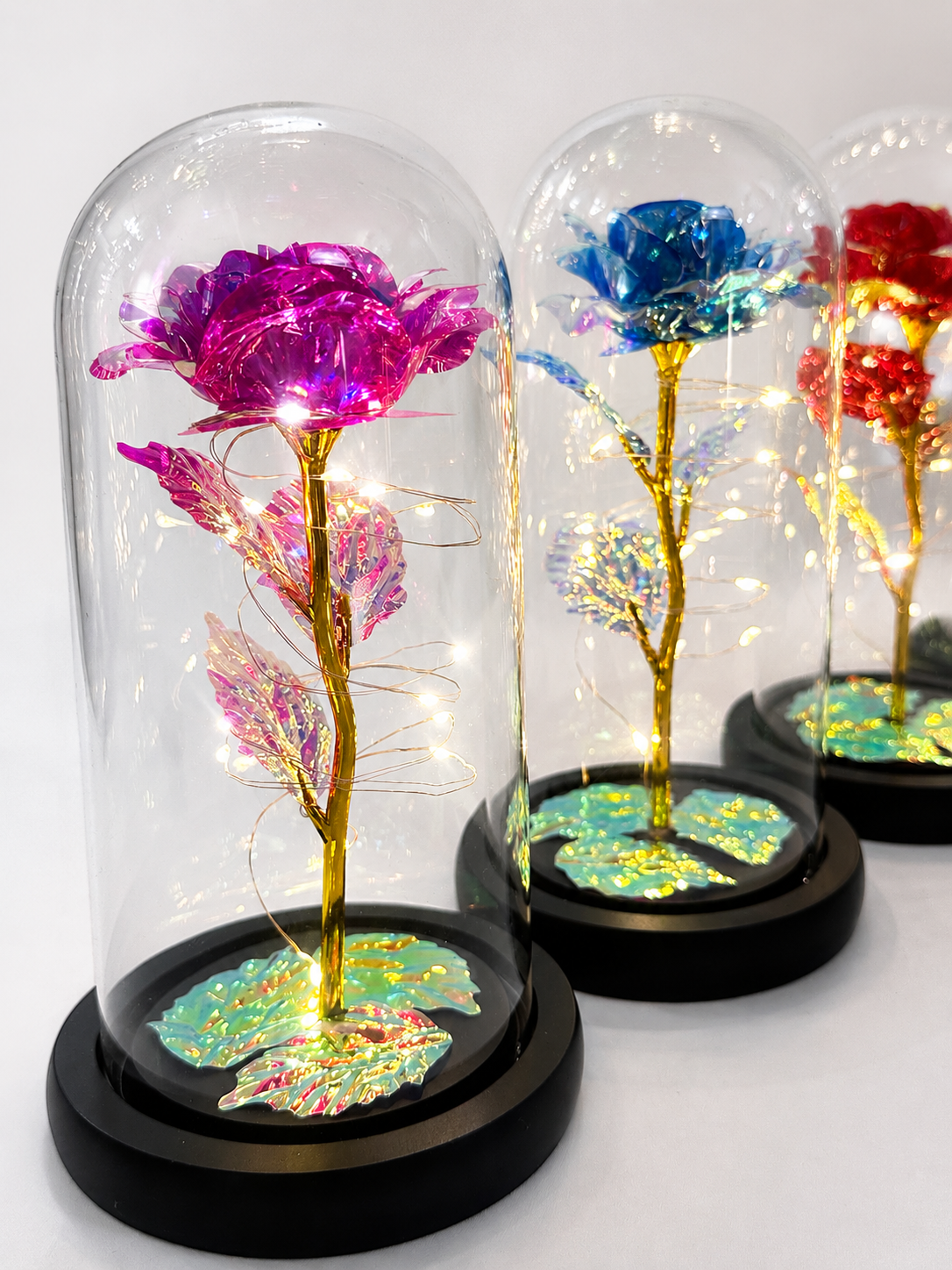 Pink blue and red close up shot of hand-made metallic roses encased in a glass dome and black wood base, three items (in different colours) are beside each other on a white background. Perfect gift-able item.