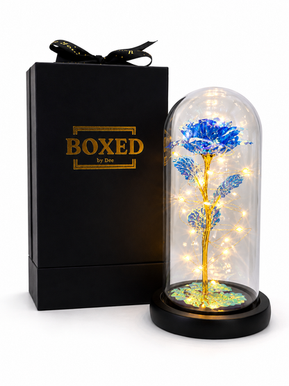 A blue metallic rose displayed under an illuminated glass dome and presented beside a beautiful Boxed by Dee gift box with a white background.