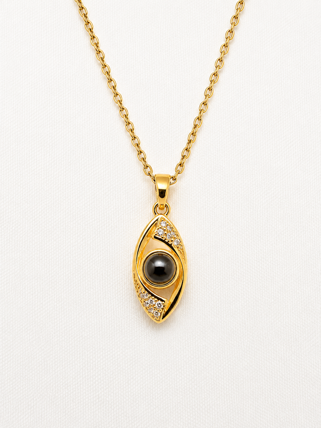 Gold evil eye pendant necklace with a black pupil and small clear stones, shown on a white textured background.