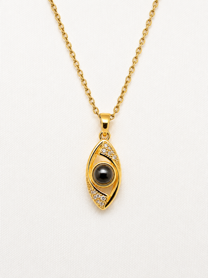 Gold evil eye pendant necklace with a black pupil and small clear stones, shown on a white textured background.