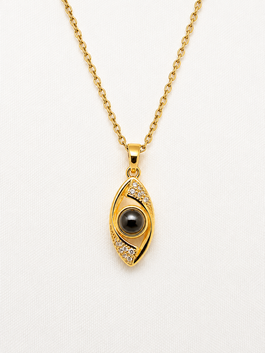 Gold evil eye pendant necklace with a black pupil and small clear stones, shown on a white textured background.