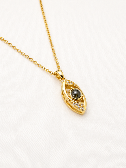 Gold evil eye pendant necklace with a dark gray pearl and small clear crystals, displayed on a textured white background.