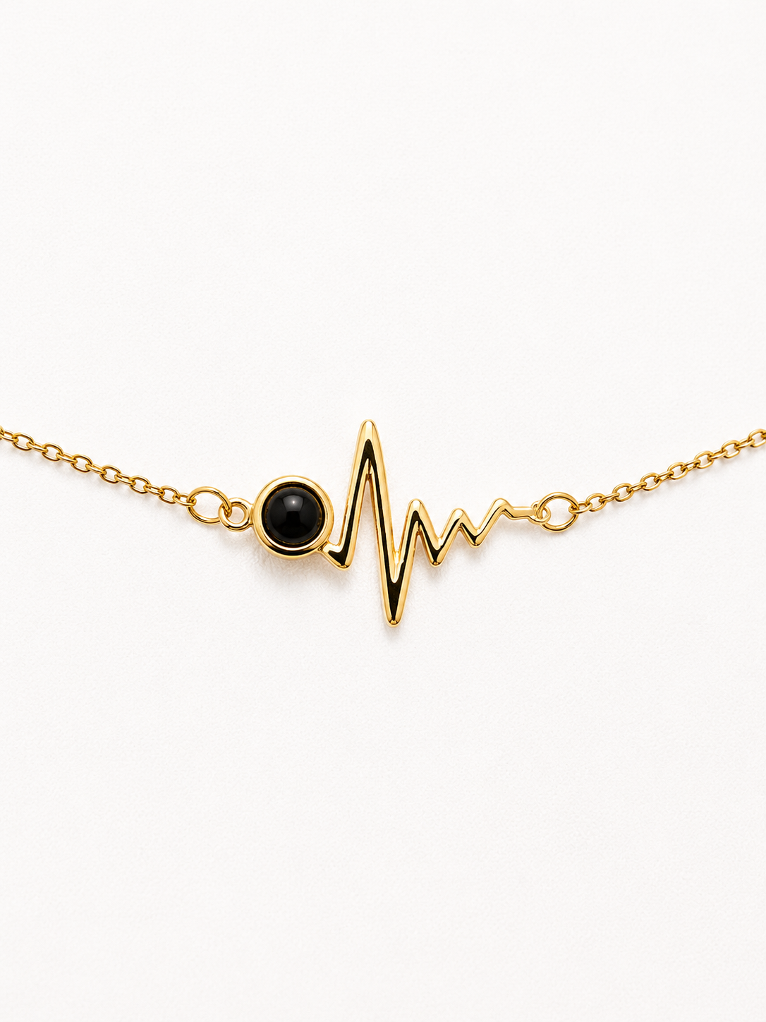 A delicate gold chain bracelet featuring a heartbeat EKG line pendant with a round black onyx stone at the heart, set against a clean white background.