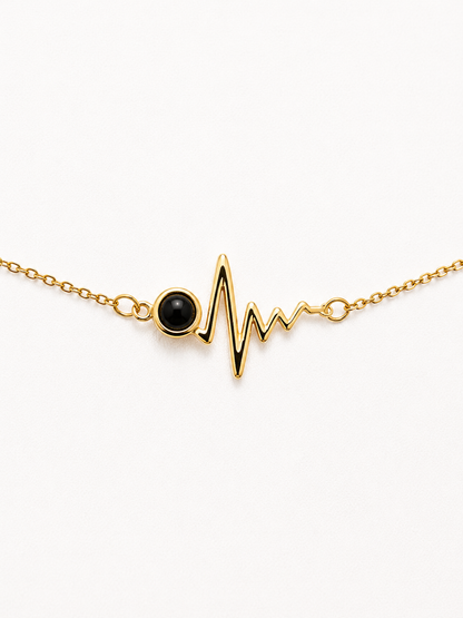 A delicate gold chain bracelet featuring a heartbeat EKG line pendant with a round black onyx stone at the heart, set against a clean white background.