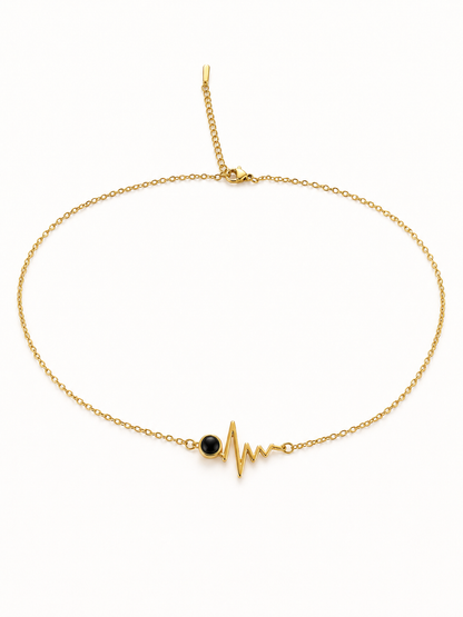 A delicate gold chain necklace featuring a central gold heartbeat EKG line charm with a small black oval stone accent, displayed on a clean white background.