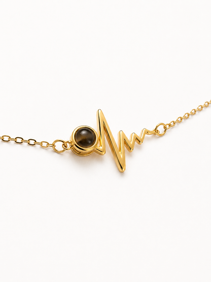 A gold chain necklace featuring a heartbeat monitor-style pendant with a small round dark brown stone, photographed on a clean white background.