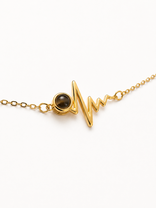 A gold chain necklace featuring a heartbeat monitor-style pendant with a small round dark brown stone, photographed on a clean white background.