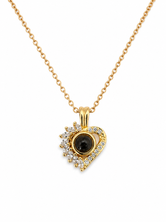 A gold heart-shaped pendant necklace / jewellery piece, featuring a central black oval stone surrounded by small clear stones, suspended from a delicate gold chain on a white background. 
