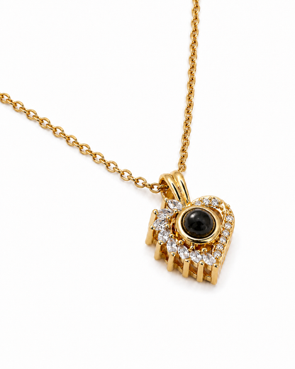 A gold pendant necklace (Jewellery piece) heart shape featuring diamantés all around with a central round black stone, shown on a white background. 