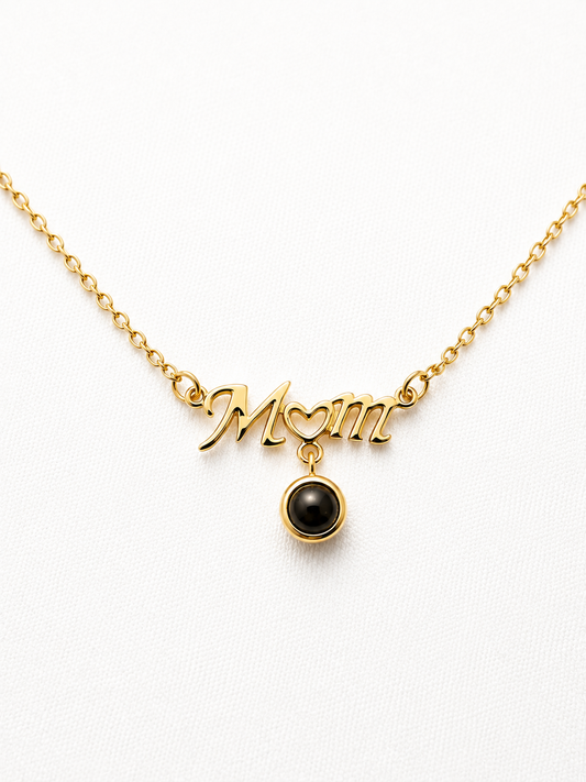 A gold chain necklace featuring a 'Mom' script pendant with a heart detail and a dangling black pearl, shown on a clean white background.