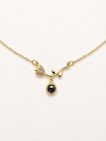 A gold chain necklace featuring a branch-shaped pendant with a small cluster of clear stones and a dangling black oval bead, displayed on a textured white background.