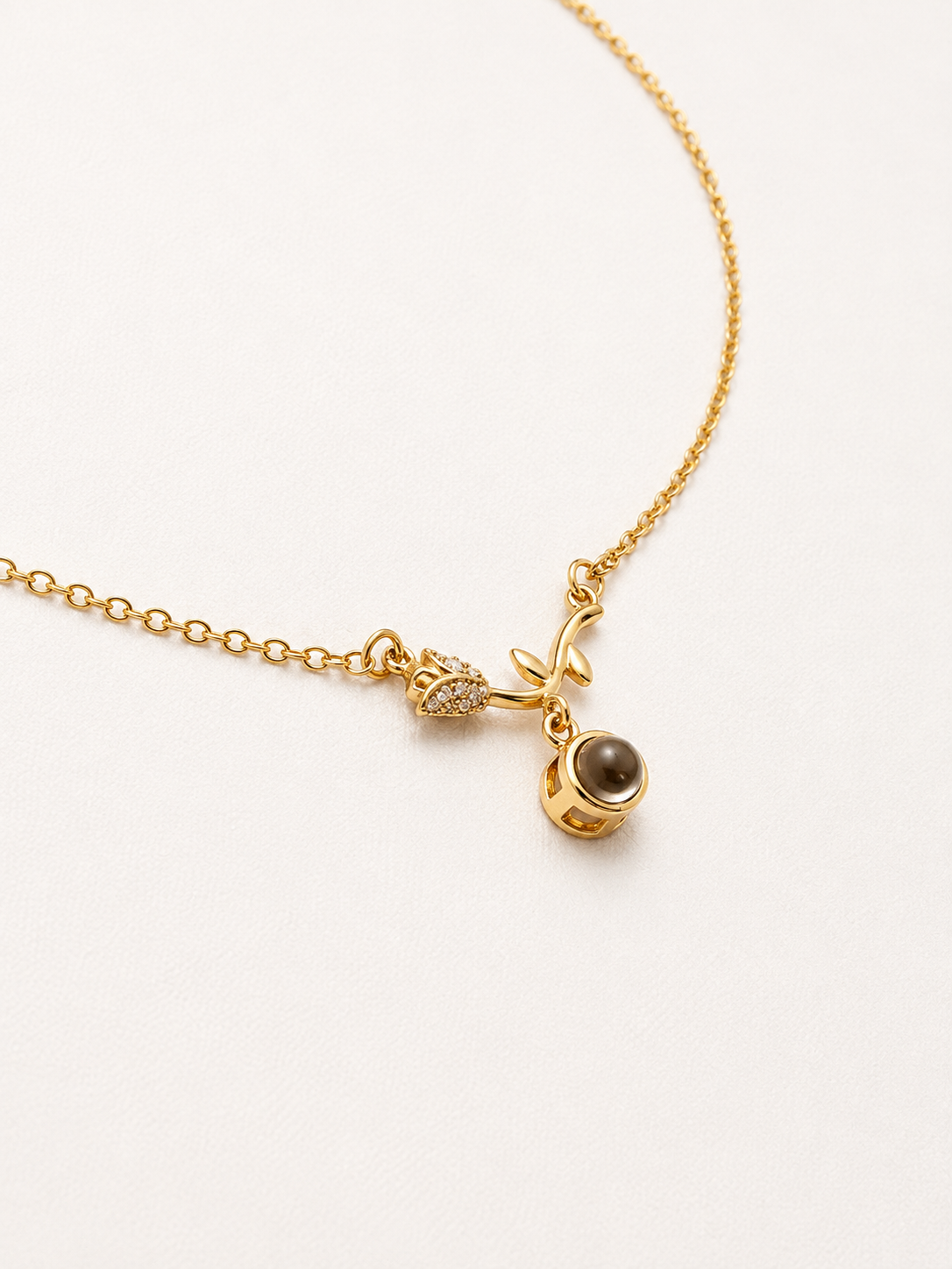 A delicate gold chain necklace featuring a small charm with a tiny clear crystal and a larger charm with a smooth, dark brown oval stone, displayed on a clean white background.