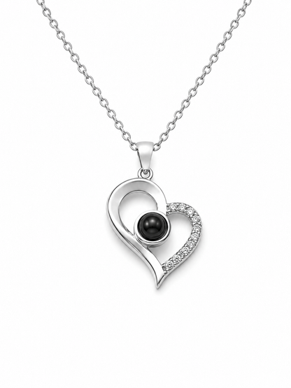 A silver heart pendant necklace featuring a polished silver heart outline with a small black pearl in the center and a pavé-set row of clear stones along the right side, suspended from a delicate silver chain against a plain white background.