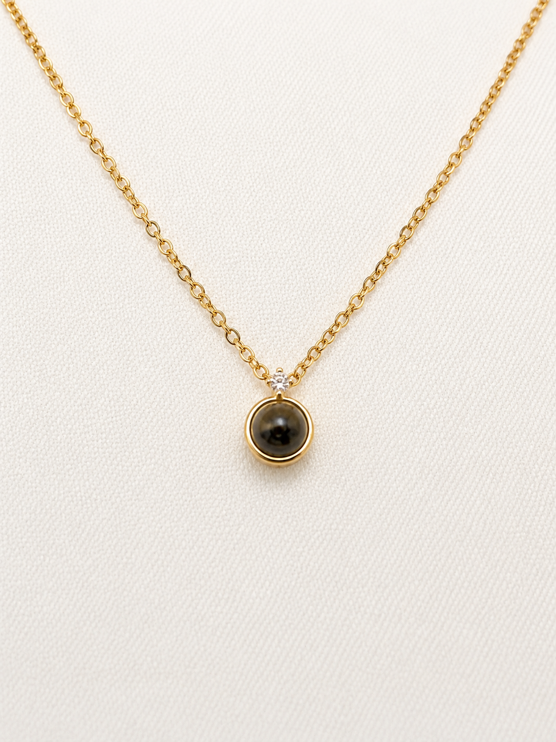 A delicate gold chain necklace with a small circular pendant featuring a dark brown tiger's eye stone and a tiny clear accent stone, displayed on a textured off-white background.