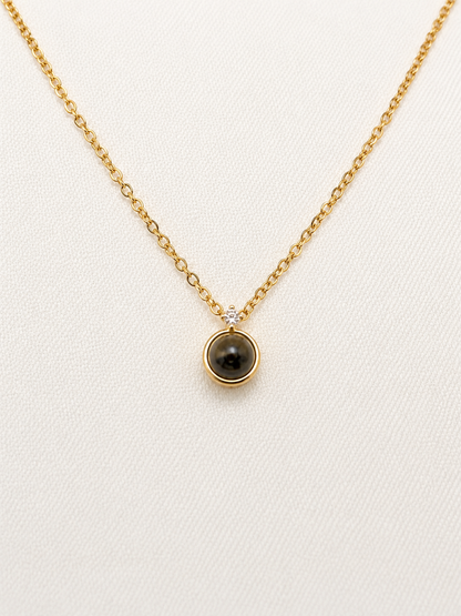 A delicate gold chain necklace with a small circular pendant featuring a dark brown tiger's eye stone and a tiny clear accent stone, displayed on a textured off-white background.