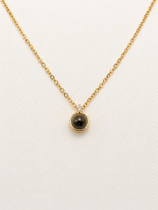 A delicate gold chain necklace with a small circular pendant featuring a dark brown tiger's eye stone and a tiny clear accent stone, displayed on a textured off-white background.