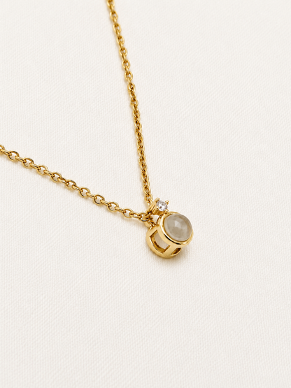 A gold chain necklace with a small pendant featuring a round pale gray stone and a tiny clear accent stone, photographed on a light cream textured background.