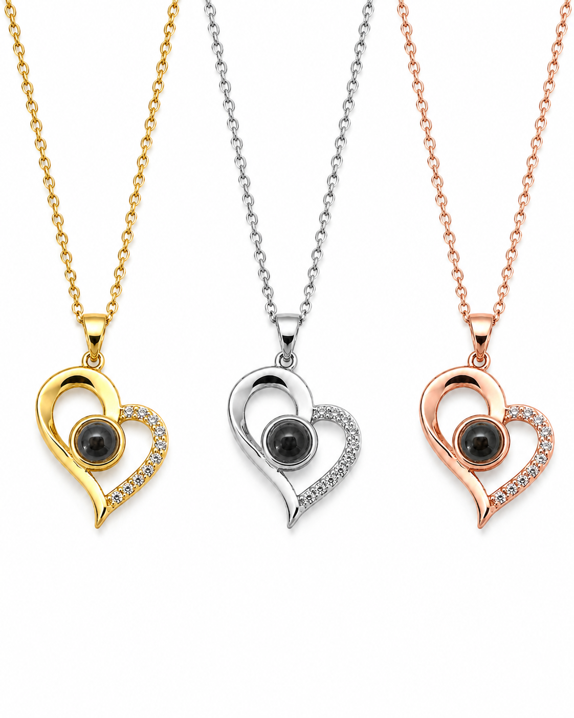 Three heart-shaped pendant necklaces shown on a white background, each with a different metal finish: yellow gold, silver, and rose gold. Each pendant features a black oval stone in the center and a curved halo of small clear stones along one side.