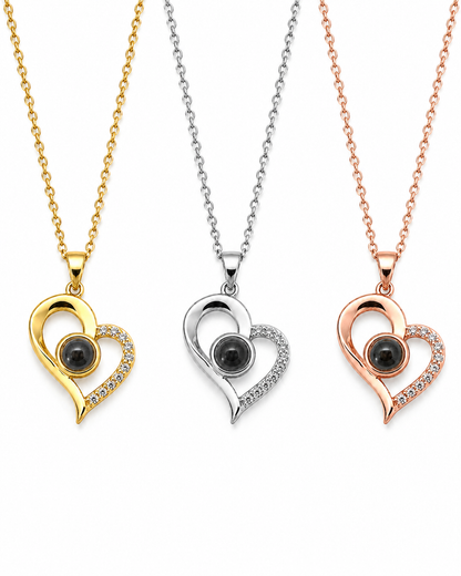 Three heart-shaped pendant necklaces shown on a white background, each with a different metal finish: yellow gold, silver, and rose gold. Each pendant features a black oval stone in the center and a curved halo of small clear stones along one side.