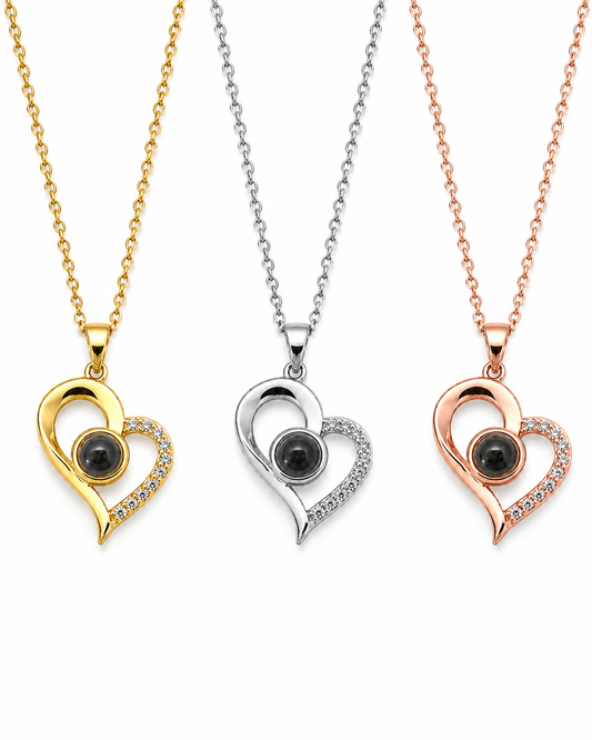 Three heart-shaped pendant necklaces shown on a white background, each with a different metal finish: yellow gold, silver, and rose gold. Each pendant features a black oval stone in the center and a curved halo of small clear stones along one side.